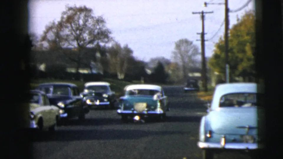 A wider home-movie view shows several mid-century cars on a Camden street, with the green-and-white sedan receding into the distance.