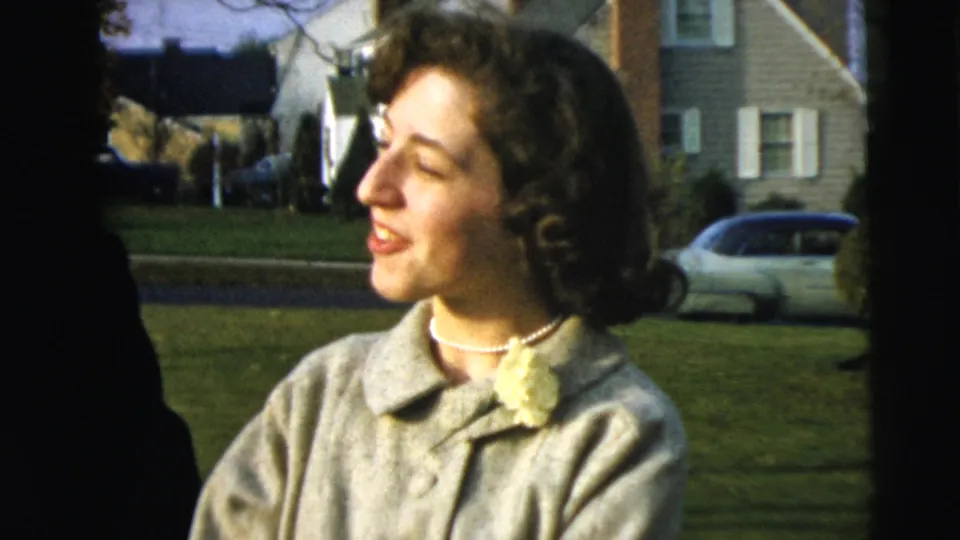 A woman in a light coat smiles outdoors in Camden, New Jersey, with a lawn, houses, and parked early 1960s cars behind her.