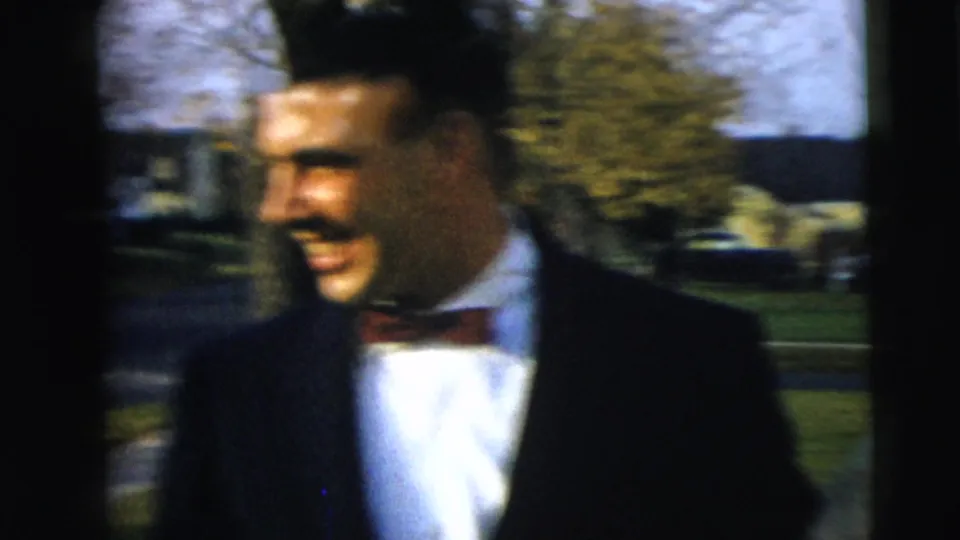 A smiling man in a dark suit and bow tie stands outside in 1962 Camden home-movie footage, with trees, lawn, and parked cars behind him.