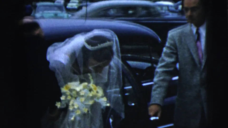 The bride in a white veil holds a yellow and white bouquet as she bends toward a dark car in the 1962 Camden wedding footage.
