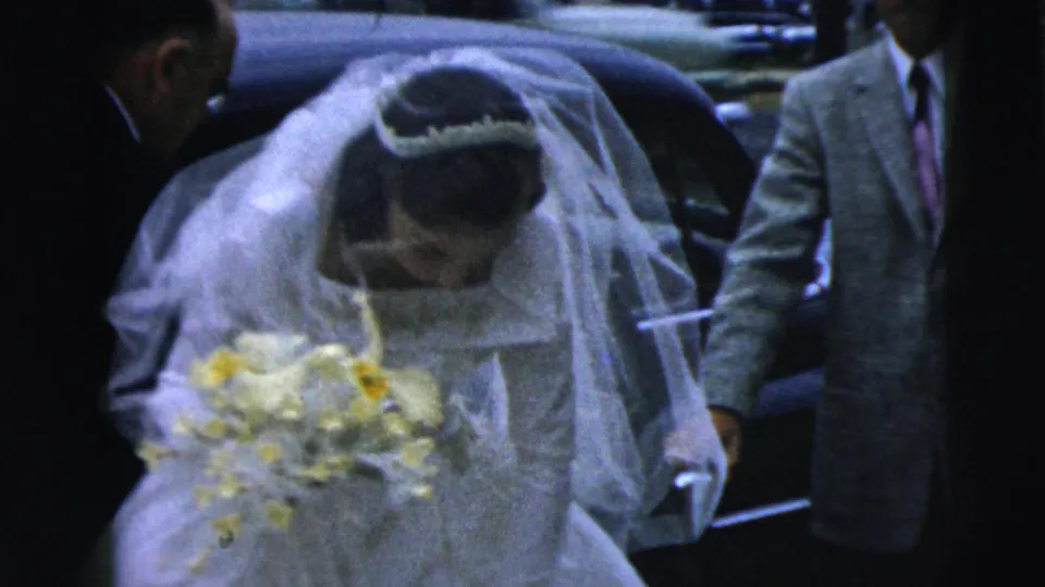 The bride, veiled and carrying a yellow-white bouquet, stands beside a car with adults nearby in Camden, New Jersey 1962 home-movie footage.
