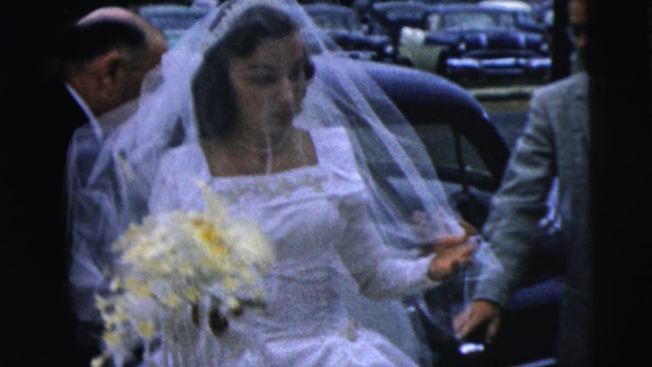 In Camden, New Jersey in 1962, a veiled bride holding flowers stands by a dark car as suited men assist her.