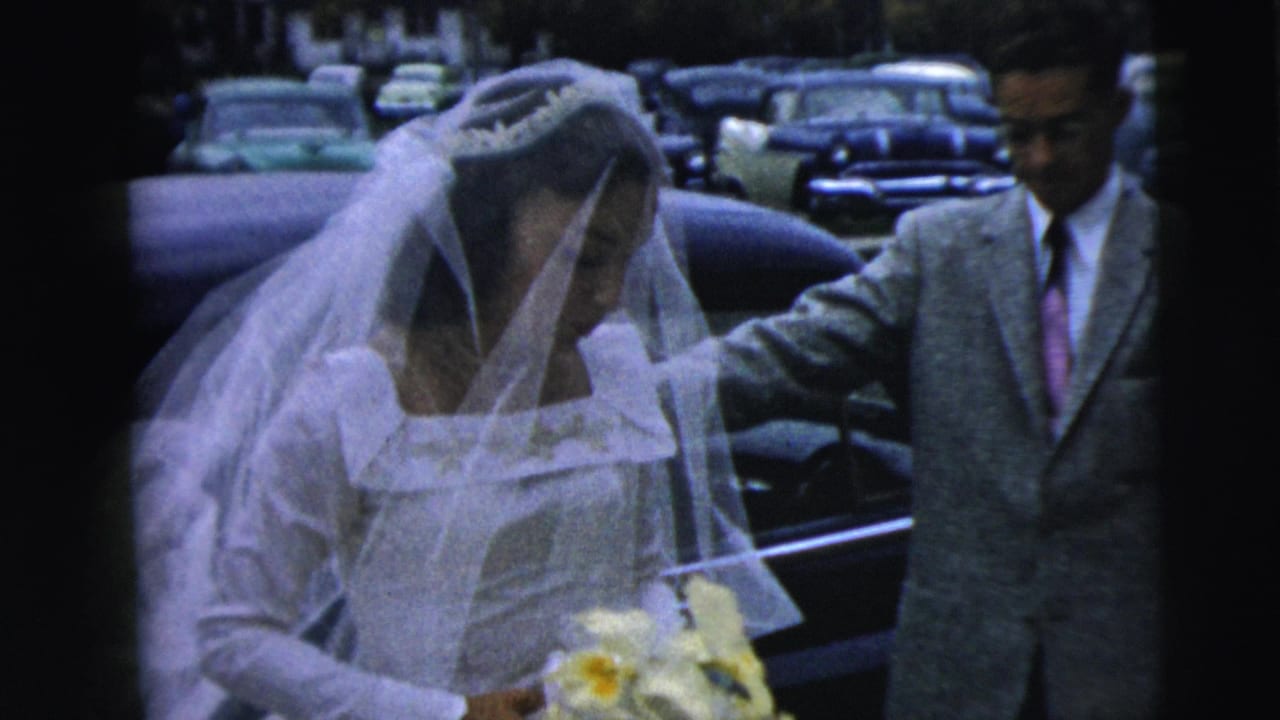 The bride in a 1962 Camden wedding scene stands beside the car while a suited man helps steady her veil or arm.