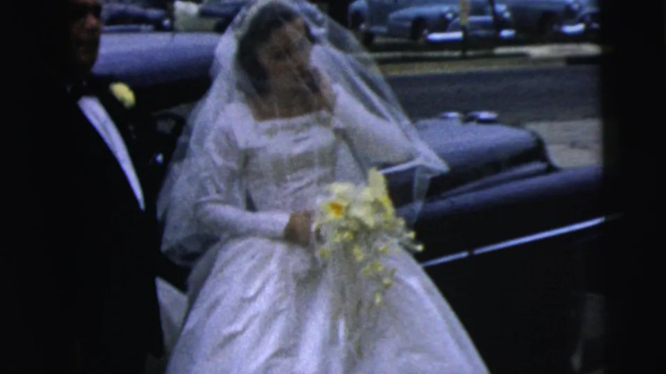 The veiled bride carries her yellow and white bouquet as she moves away from the car in the Camden wedding footage.