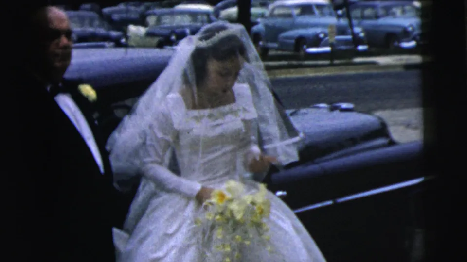 The bride pauses near the dark car during the 1962 Camden wedding arrival, bouquet in hand and suited attendant beside her.