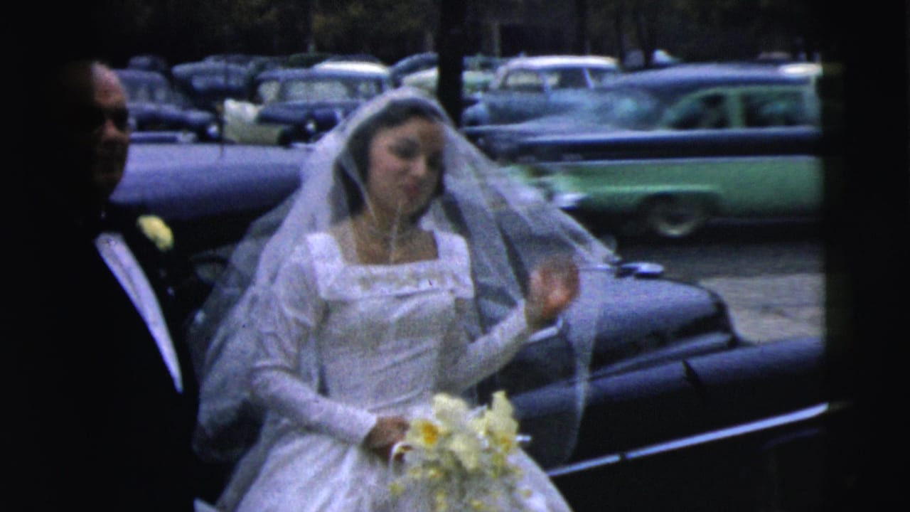 In Camden, New Jersey, 1962, a bride in a white gown and veil stands beside a limousine as a tuxedoed man remains at her side.