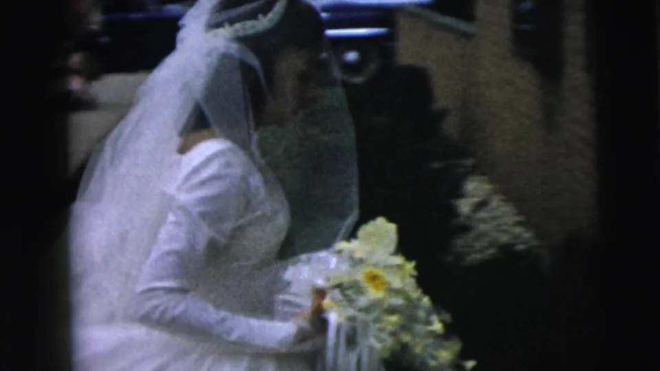 The bride leans forward near the limousine, holding flowers while another person appears to help manage her gown or veil.