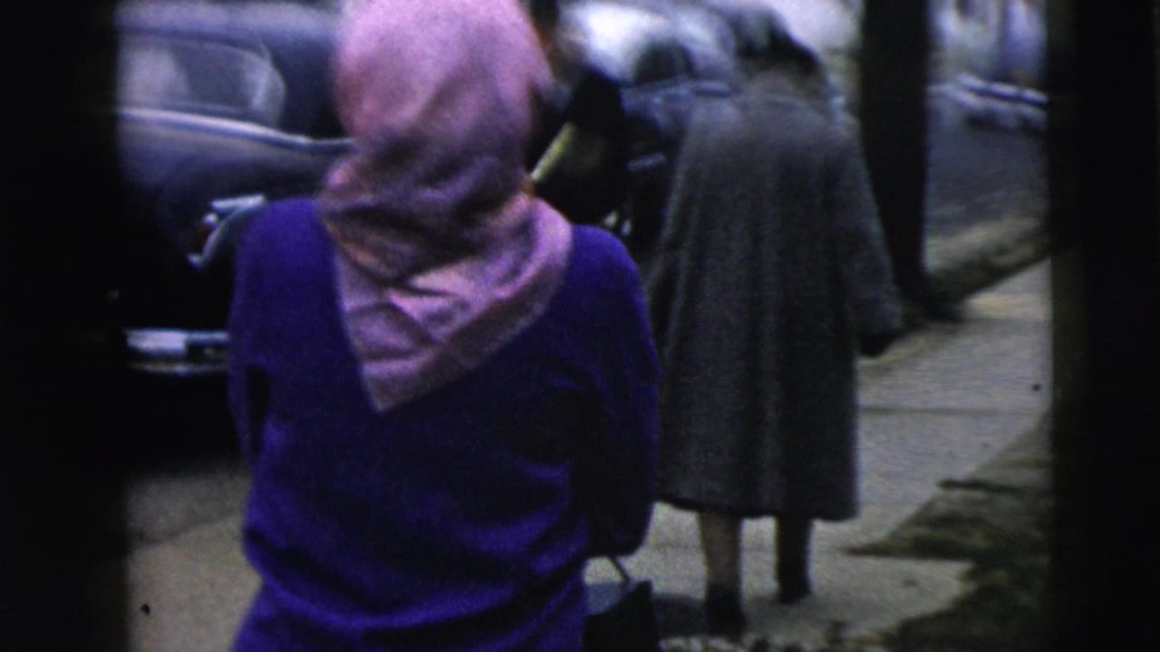 In Camden, New Jersey, a woman in a purple sweater and pink headscarf walks along a sidewalk near parked classic cars and pedestrians.