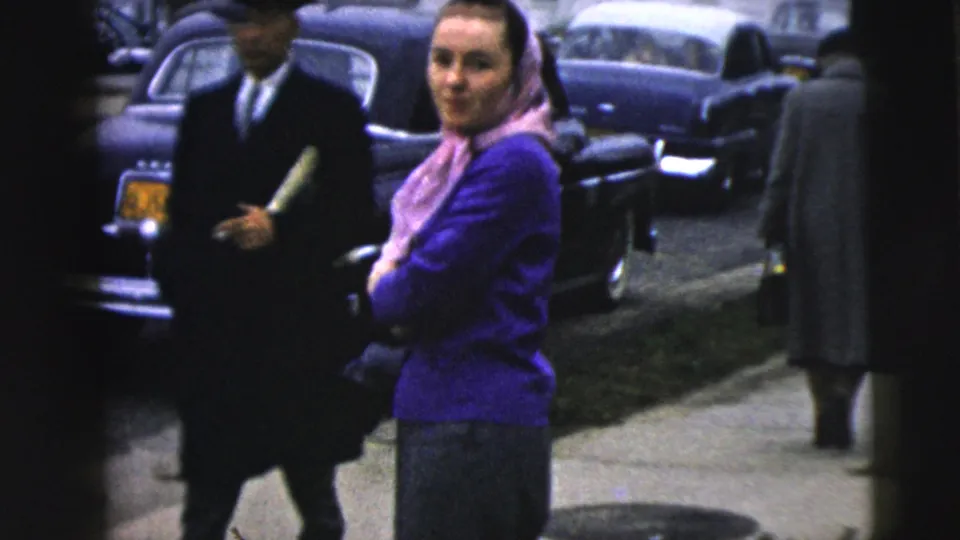 In a 1962 Camden street scene, a woman in a purple sweater and pink headscarf looks toward the camera beside classic cars and passing pedestrians.