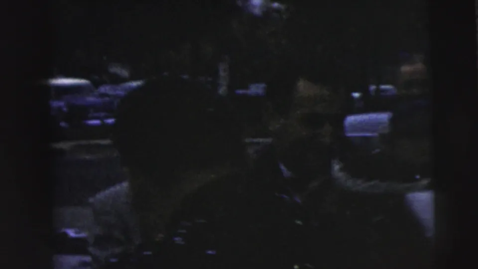 Low-exposure 1962 Camden wedding departure frame showing blurred figures near or inside a car, with seats and reflective highlights visible.