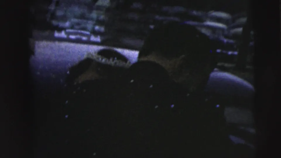 Dark 1962 Camden home-movie frame showing an adult close to a long gun in a wooded outdoor setting.