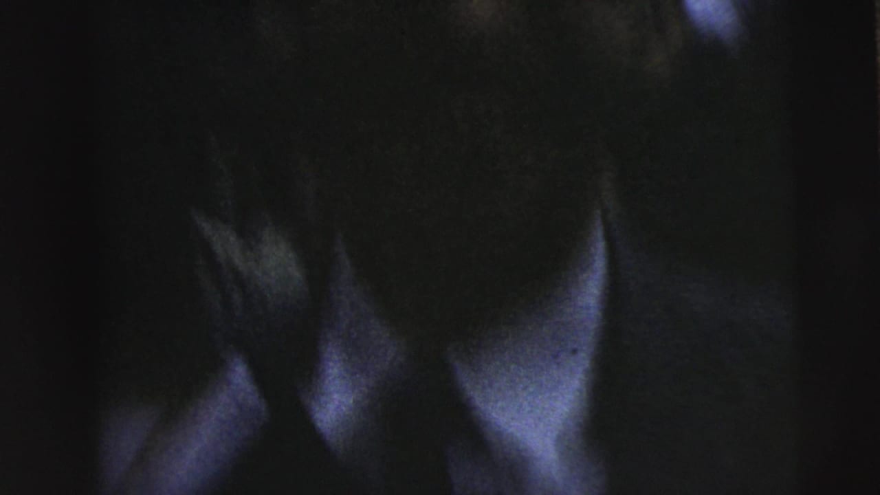 Very dark 8mm frame from the Camden clip with a close-up of an adult face, shirt collar, and jacket edge.