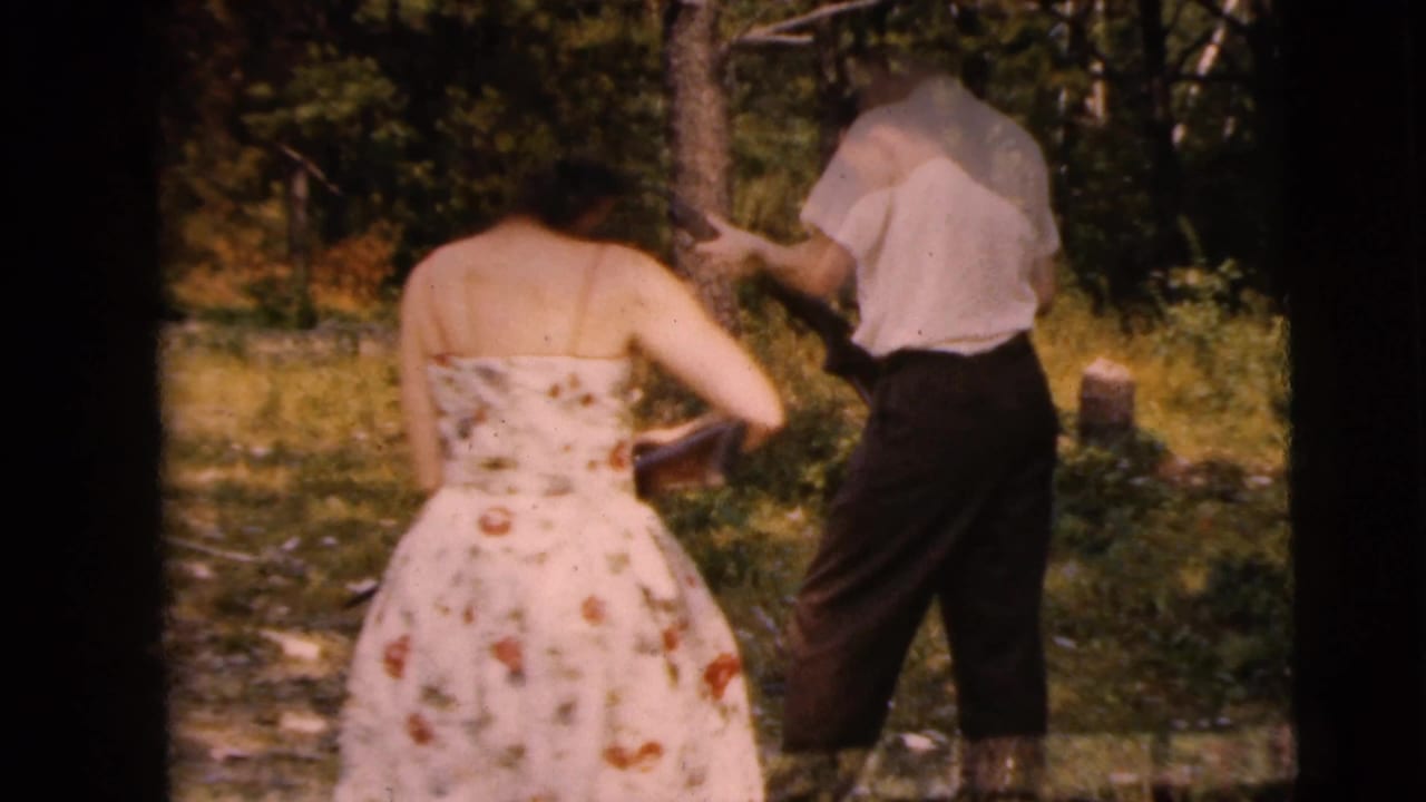 Woman in floral strapless dress and man in white shirt stand in wooded grass with rifles nearby.