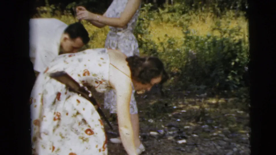 In this 1962 wooded home-movie frame, a woman in a floral dress bends down with a rifle visible near her side as others stand nearby.
