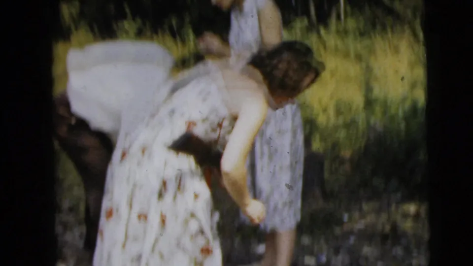 A soft, motion-blurred 1962 Camden-area home-movie frame shows a woman bending with a rifle while another woman stands nearby in tall grass.