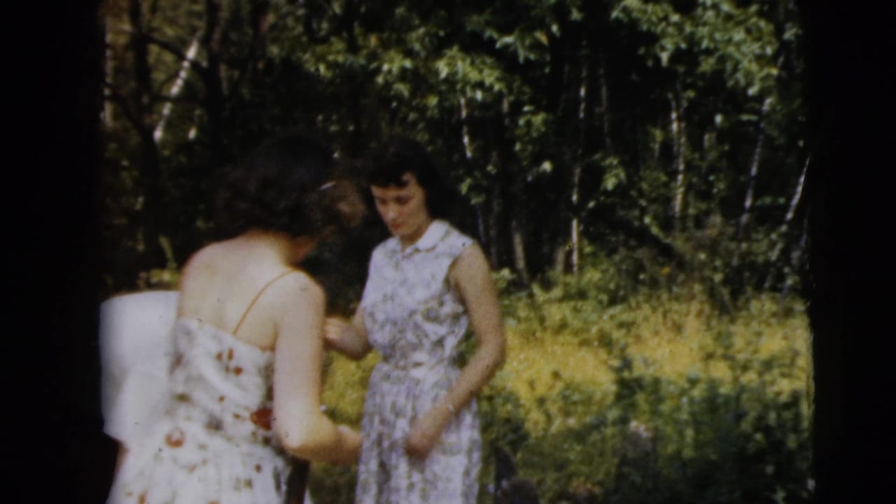 Two adult women in patterned dresses stand in a wooded area near a long gun.