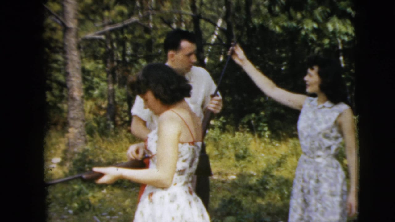 Adults in a wooded clearing handle long guns while one woman gestures upward.