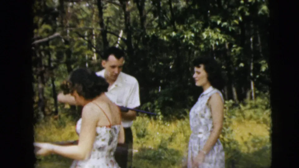 In the Camden-area 1962 8mm footage, a woman holds a long gun as a man in a white shirt and another woman stand behind her in the trees.