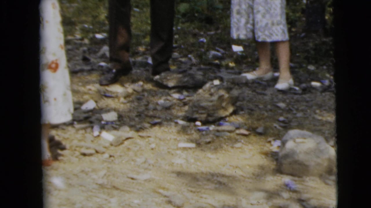 Adults' lower bodies stand on a dirt path in woods with scattered debris and trees nearby.