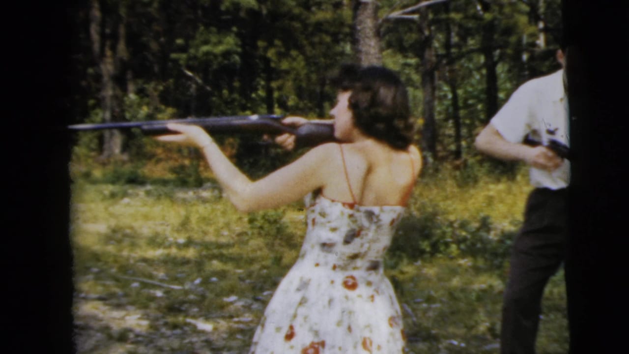 1962 Camden 8mm frame of a woman in a floral dress aiming a rifle in woods, with a man in a white shirt nearby.