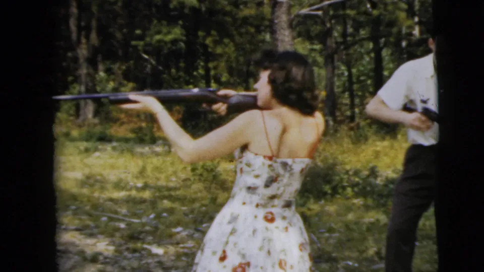 1962 Camden 8mm frame of a woman in a floral dress aiming a rifle in woods, with a man in a white shirt nearby.