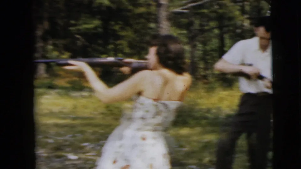 1962 Camden home-movie frame showing a woman aiming a rifle in woods with a man holding another firearm in the background.