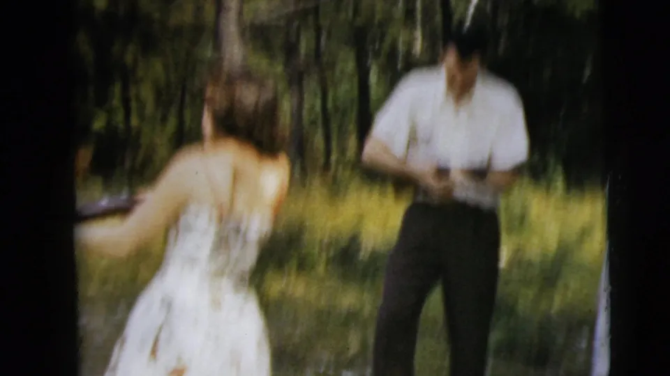 Blurred 1962 Camden 8mm frame with a woman seen from behind and a man holding a firearm in a wooded clearing.