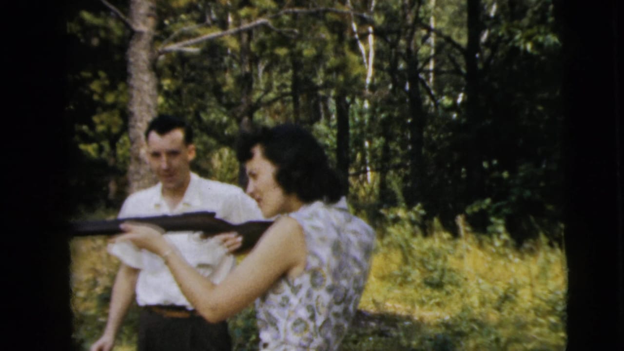 In 1962 Camden, New Jersey home-movie footage, a woman in a patterned sleeveless dress aims a rifle as a man watches in a wooded area.