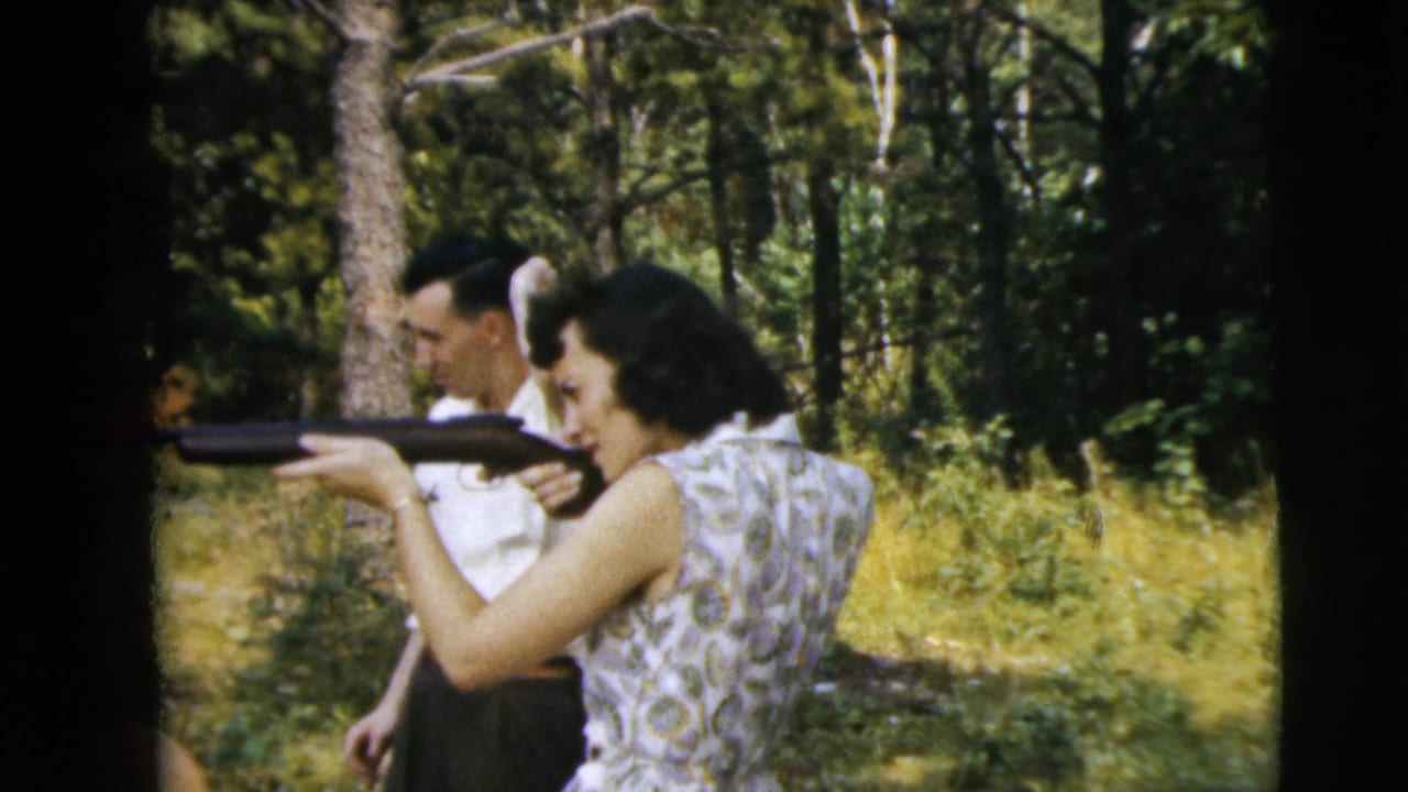 A woman shoulders a rifle in a sunlit wooded area, with a man in a white shirt observing behind her in 1962 home-movie footage.