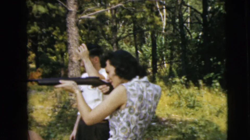 The woman keeps the rifle aimed in the woods while the man behind her raises an arm, captured with soft 8mm color grain.