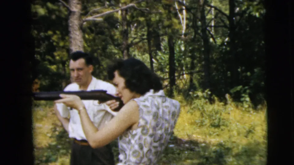 A woman in a patterned dress aims a rifle in the foreground while a man watches from a wooded clearing in Camden, New Jersey metadata.