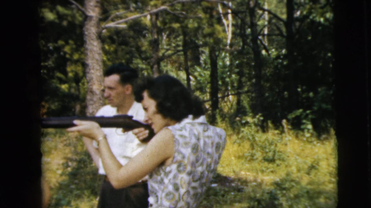 Woman aiming a long gun outdoors while a man stands behind her among trees.