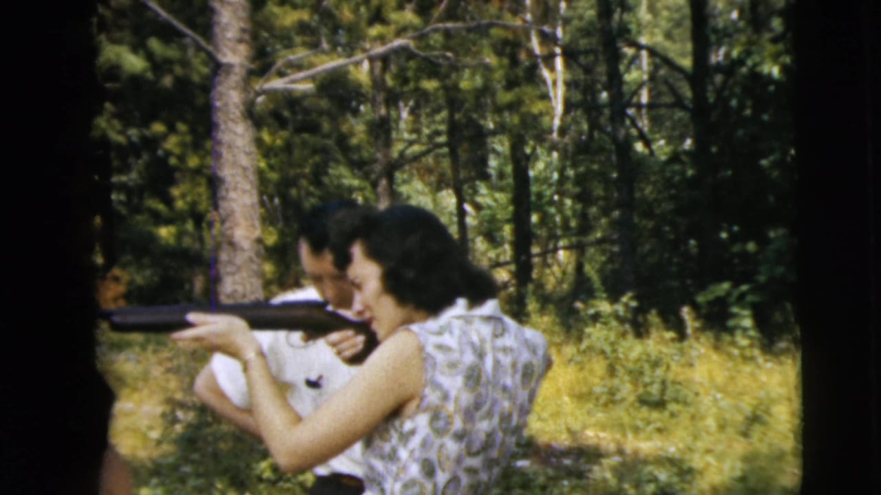Man and woman in a wooded yard aim long guns toward the left.