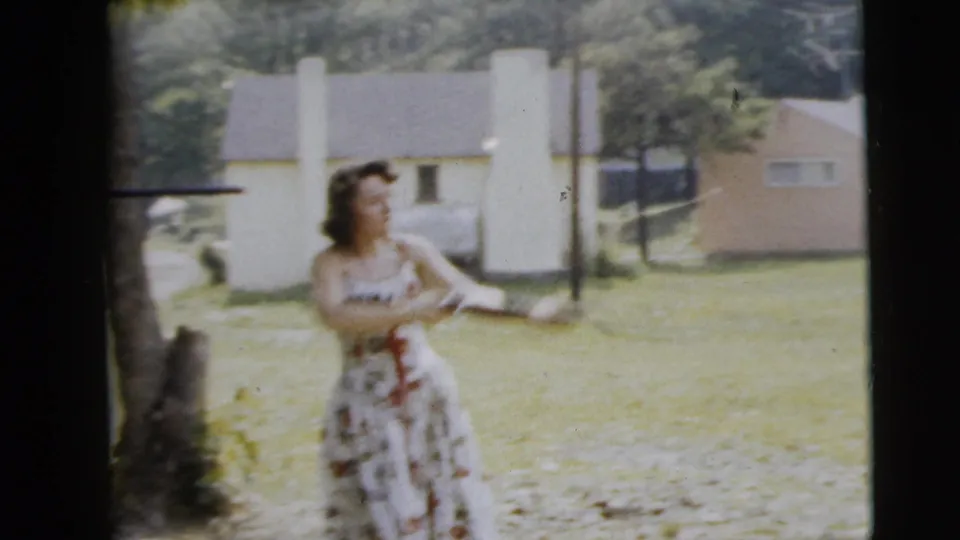 A woman in a sleeveless patterned dress handles a long gun in a grassy yard, with a house and trees visible behind her.
