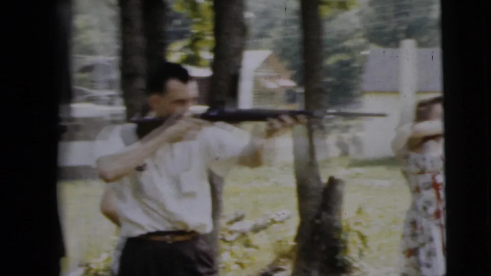 A man in a white shirt aims a long gun in a grassy yard, with trees, a house, and a partially visible woman nearby.