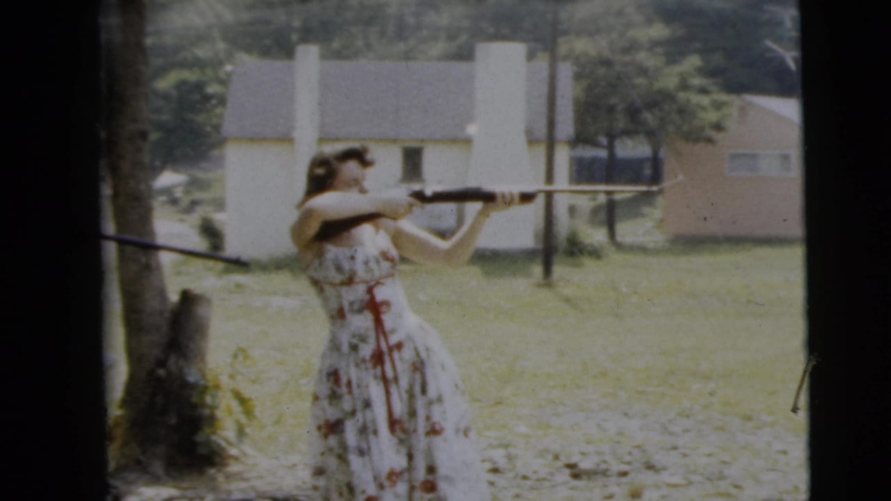 In 1962 Camden, New Jersey home-movie footage, a woman in a white floral dress aims a shotgun in a sunlit yard with houses behind her.