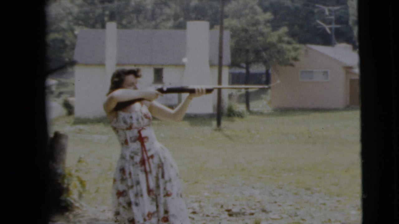 The woman keeps the shotgun raised while standing outdoors in the Camden, New Jersey yard, with pale houses and trees visible beyond her.