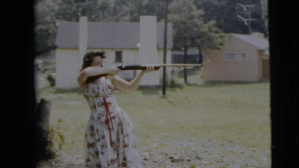 The woman keeps the shotgun raised while standing outdoors in the Camden, New Jersey yard, with pale houses and trees visible beyond her.