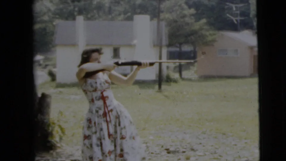 A soft 8mm frame shows the woman in a patterned floral dress aiming the shotgun, with a tree trunk, grass, and suburban houses behind her.