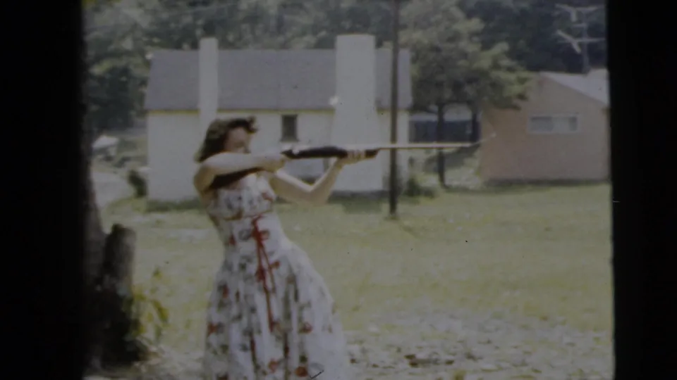 The final frame keeps the woman and shotgun centered against the bright yard, with houses, trees, and visible 8mm frame edges in view.