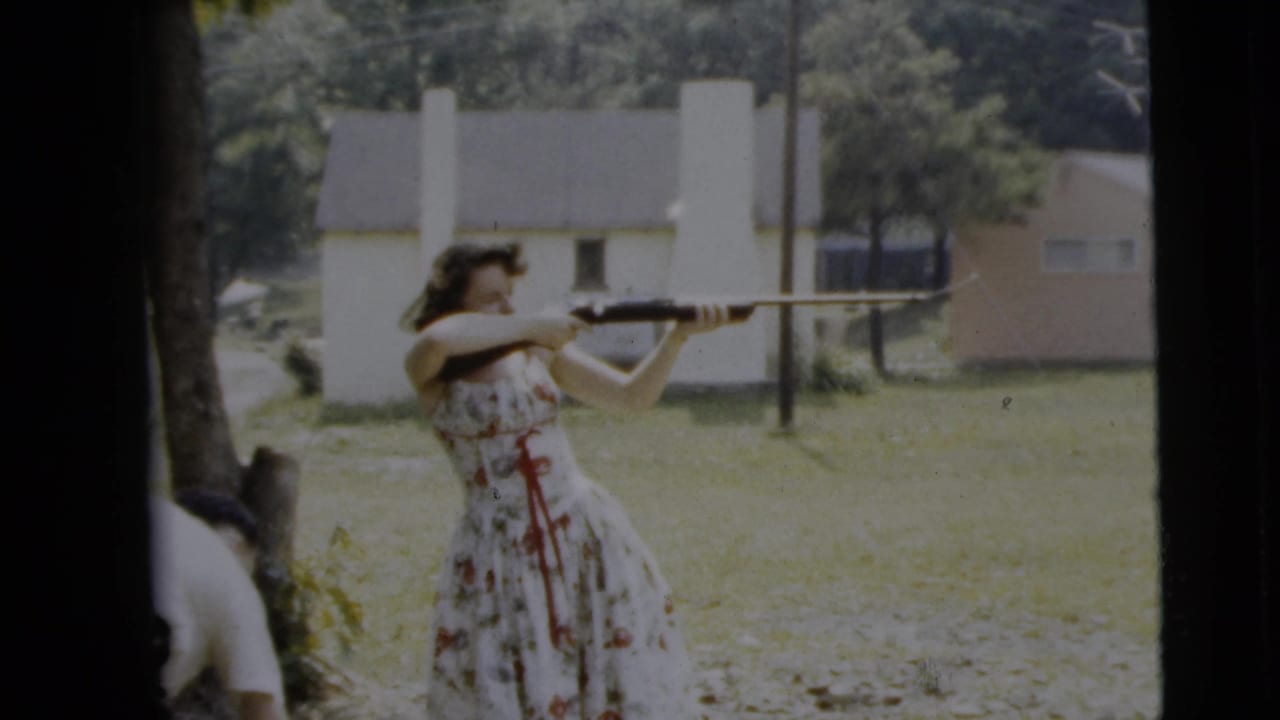 Woman in a floral dress aims a long rifle in a grassy yard with houses behind her.