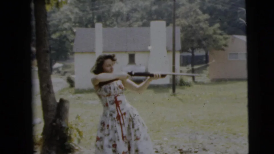 A woman in a floral dress keeps a rifle raised in a Camden backyard scene dated 1962, with houses and trees in the background.