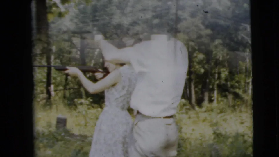 A man in a white shirt stands close to a woman with a rifle during a 1962 Camden backyard lesson, partly softened by exposure and film grain.