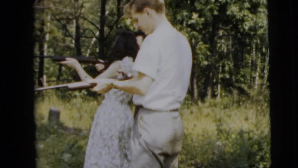 The man appears to guide the woman's stance as she holds the rifle in a grassy Camden yard, captured in 1962 8mm home-movie color.