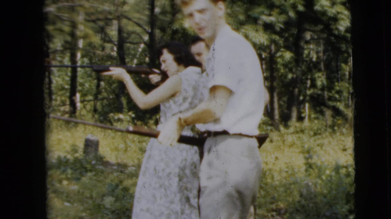 Three adults in woods as a woman aims a rifle with assistance from two men.