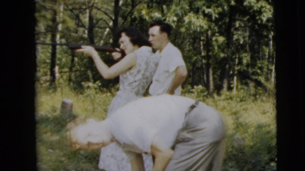 Woman aims a rifle while one man stands behind her and another bends forward nearby.