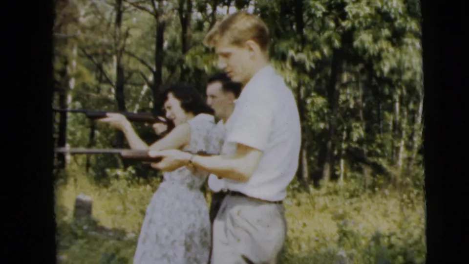 Two men and a woman stand close together in 1962 wooded home-movie footage, with a rifle held across the foreground and another aimed left.