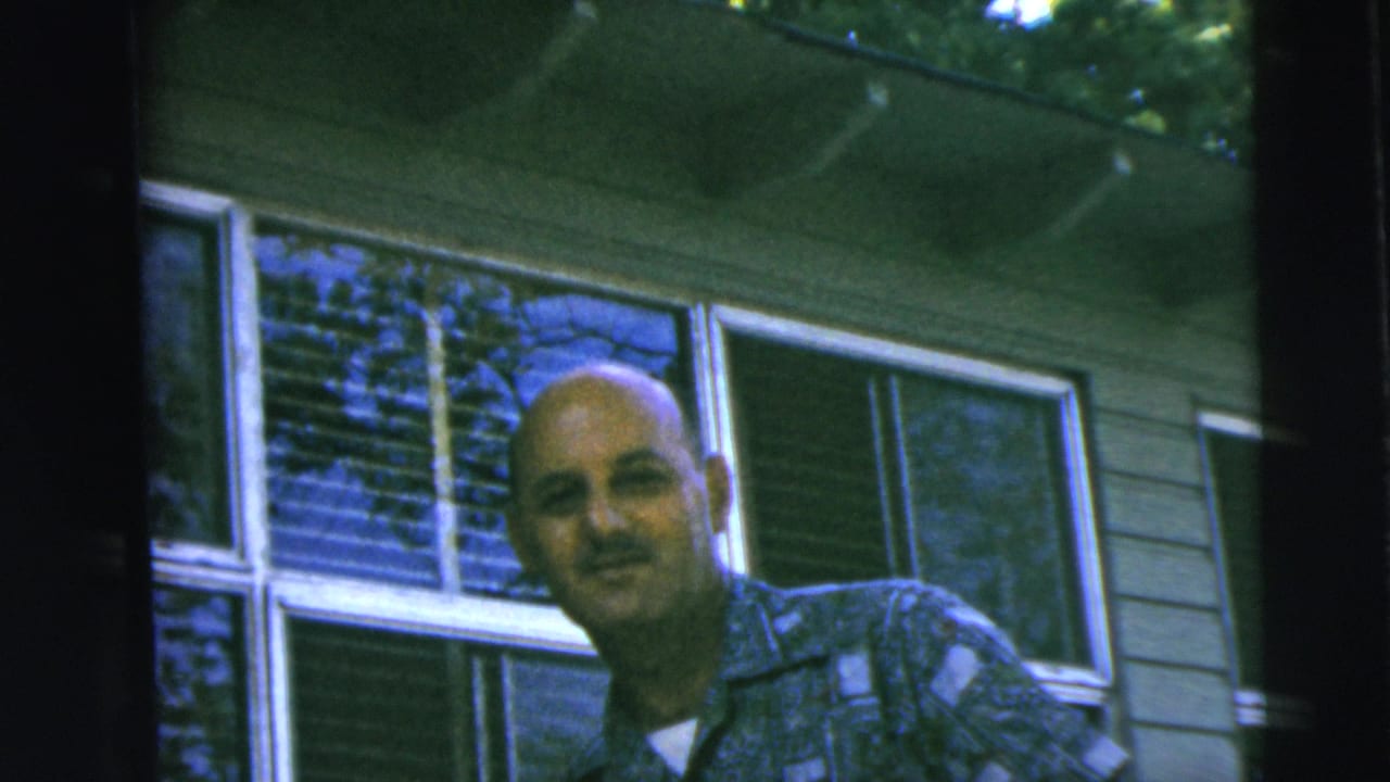 A bald adult man smiles toward the camera outside a Camden, New Jersey home in the stated 1962 family footage.