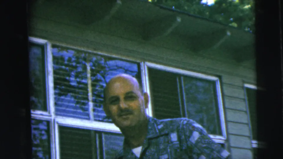 A bald adult man smiles toward the camera outside a Camden, New Jersey home in the stated 1962 family footage.