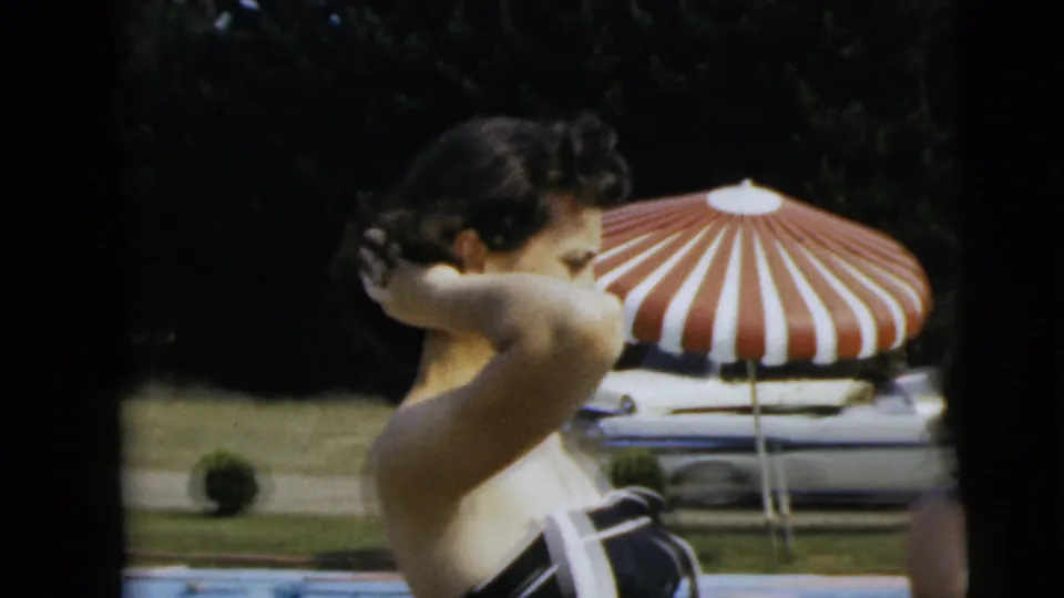 A woman in a black-and-white swimsuit adjusts her hair near a backyard pool, with a striped patio umbrella and parked car behind her.